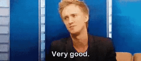 A Man In A Black Jacket Is Sitting In Front Of A Blue Wall And Saying `` Very Good '' . GIF