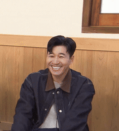 A Man In A Black Jacket Is Smiling In Front Of A Wooden Wall . GIF
