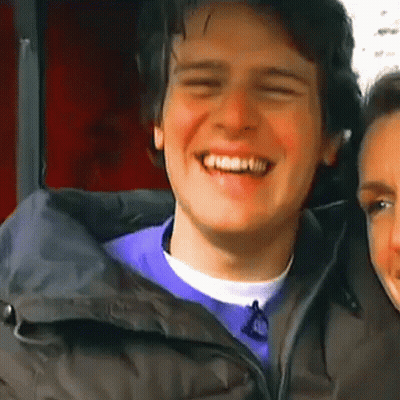 A Man In A Black Jacket Is Smiling While Standing Next To A Woman In A Purple Shirt . GIF