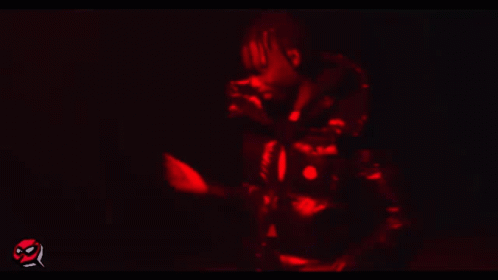 A Man In A Black Jacket Is Standing In A Dark Room With A Red Light Behind Him GIF