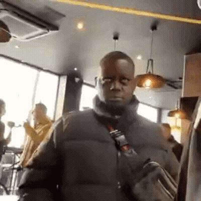 A Man In A Black Jacket Is Standing In A Restaurant With A Bag On His Shoulder . GIF