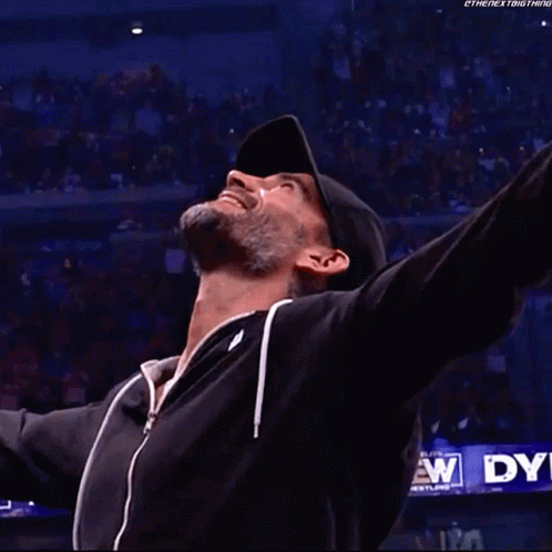 A Man In A Black Jacket Is Standing In A Wrestling Ring With His Arms Outstretched . GIF