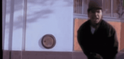 A Man In A Black Jacket Is Standing In Front Of A Clock On A Wall . GIF