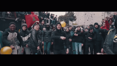 A Man In A Black Jacket Is Standing In Front Of A Crowd Of People GIF