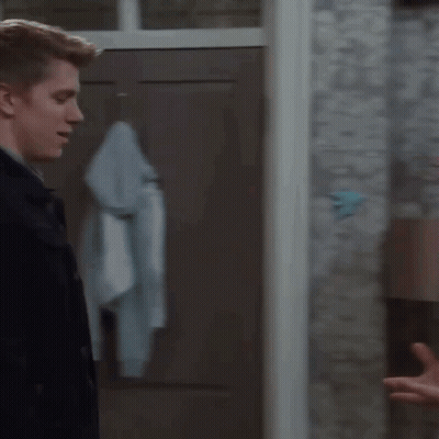 A Man In A Black Jacket Is Standing In Front Of A Door Talking To A Woman . GIF