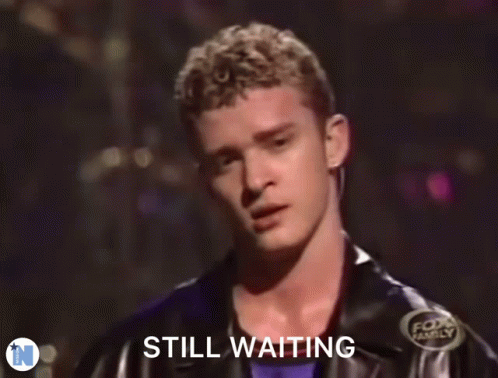 A Man In A Black Jacket Is Standing In Front Of A Sign That Says " Still Waiting " GIF