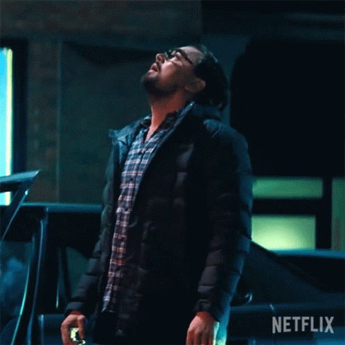 A Man In A Black Jacket Is Standing Next To A Car And Looking Up At The Sky . GIF