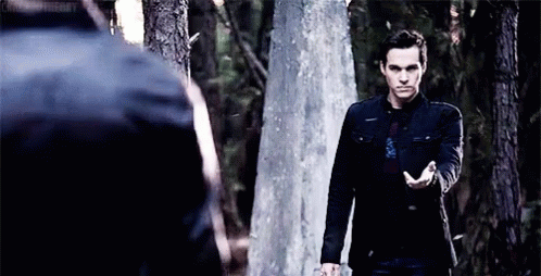 A Man In A Black Jacket Is Standing Next To A Tree In The Woods And Holding His Hand Out . GIF