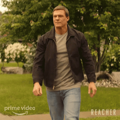 A Man In A Black Jacket Is Walking Down A Sidewalk In Front Of A Lush Green Field . GIF