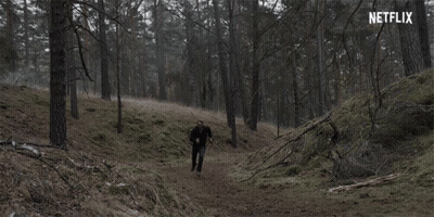A Man In A Black Jacket Is Walking Through A Dark Forest With A Netflix Logo In The Background GIF