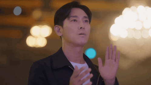 A Man In A Black Jacket Is Waving His Hand In Front Of A Chandelier GIF