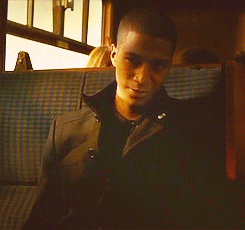 A Man In A Black Jacket Sits In A Blue Plaid Seat On A Train GIF