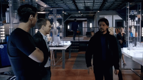 A Man In A Black Jacket Stands In A Room With Other Men And A Woman GIF