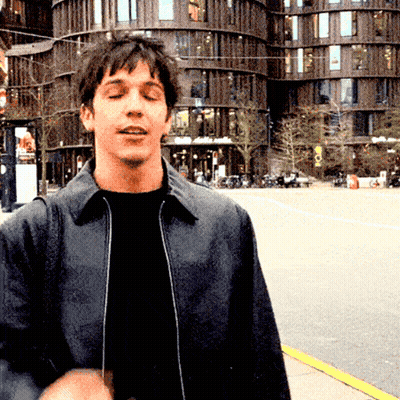 A Man In A Black Jacket Stands In Front Of A Building That Says ' A ' On The Front GIF
