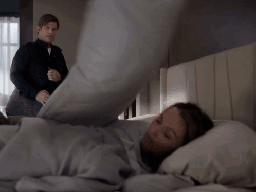 A Man In A Black Jacket Stands Next To A Man Laying In Bed GIF