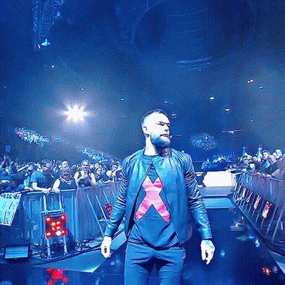 A Man In A Black Jacket With A Red X On His Shirt Stands On A Stage GIF