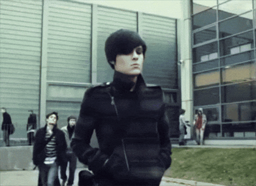 A Man In A Black Jacket With His Hands In His Pockets Stands In Front Of A Building GIF