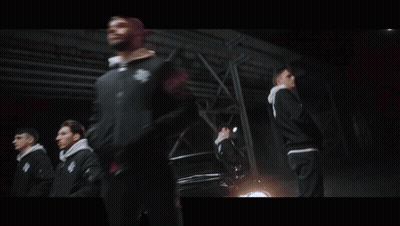 A Man In A Black Jacket With The Letter S On It Stands In Front Of A Car GIF