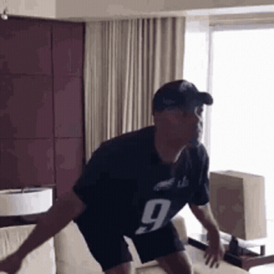 A Man In A Black Jersey With The Number 9 On It Is Jumping On A Couch In A Living Room . GIF