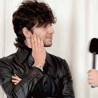 A Man In A Black Leather Jacket Is Holding His Hand To His Face While Talking Into A Microphone . GIF