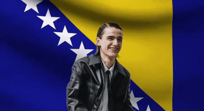 A Man In A Black Leather Jacket Is Smiling In Front Of A Flag GIF