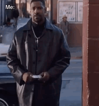 A Man In A Black Leather Jacket Is Standing On A Sidewalk Holding A Cell Phone . GIF
