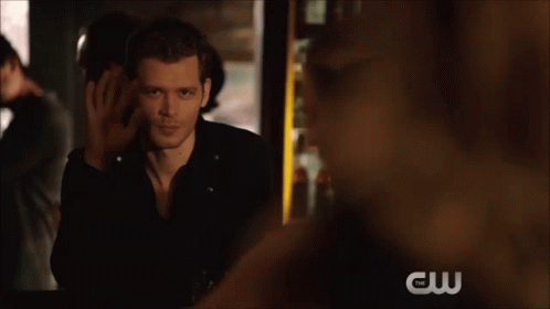 A Man In A Black Leather Jacket Sits With His Feet Up And A Cw Logo In The Background GIF