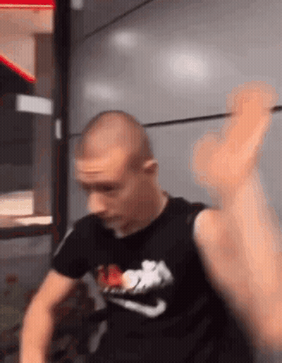A Man In A Black Nike Shirt Is Standing In Front Of A Wall With His Hand Up . GIF