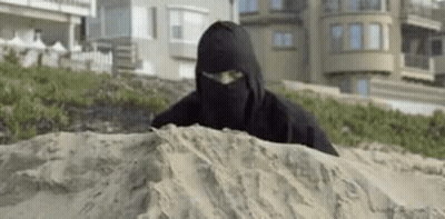 A Man In A Black Ninja Costume Is Hiding In The Sand On The Beach . GIF
