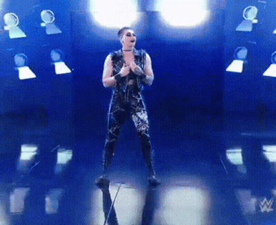 A Man In A Black Outfit Is Standing In Front Of A Wrestling Logo GIF