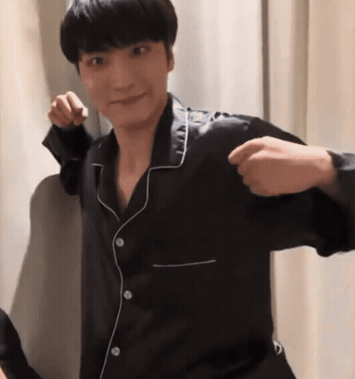 A Man In A Black Pajama Shirt Is Standing In Front Of A White Curtain And Making A Fist . GIF