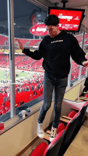 A Man In A Black Pleasures Hoodie Stands In A Stadium Watching A Football Game GIF