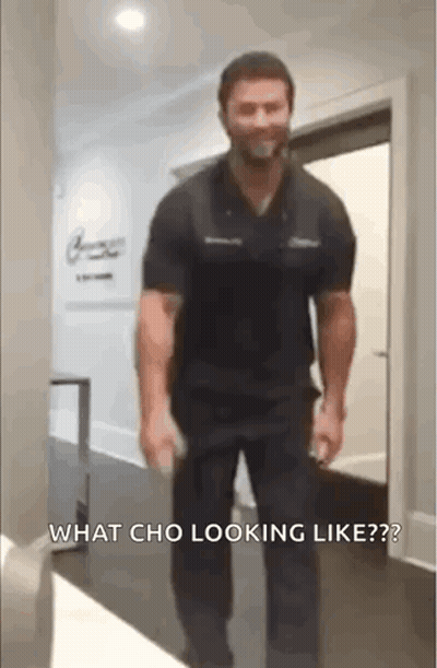A Man In A Black Shirt And Black Pants Is Walking Down A Hallway With A Bottle In His Hand . GIF