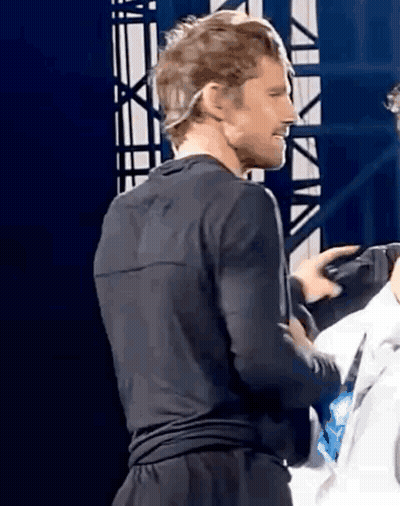 A Man In A Black Shirt And Black Shorts Is Standing On A Stage With A Microphone On His Head . GIF