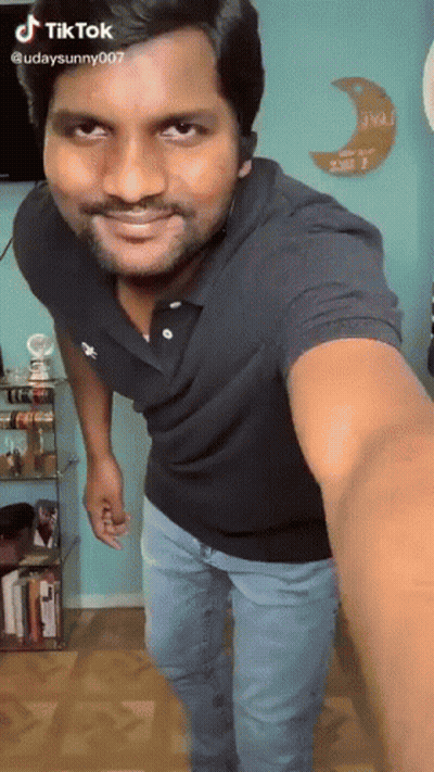 A Man In A Black Shirt And Blue Jeans Is Taking A Selfie In A Room . GIF