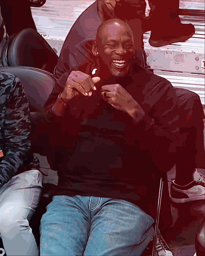 A Man In A Black Shirt And Blue Jeans Sits In A Chair And Laughs GIF