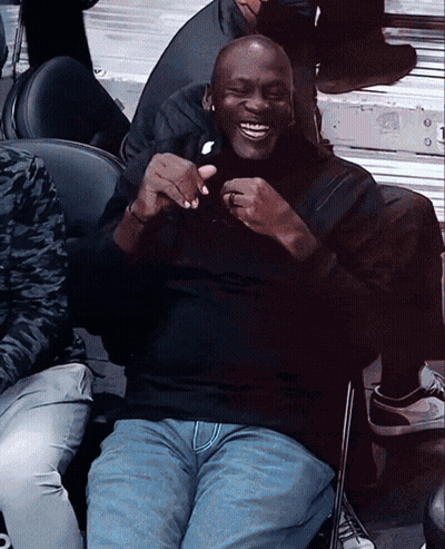 A Man In A Black Shirt And Blue Jeans Sits In A Chair And Smiles GIF