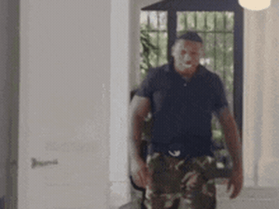 A Man In A Black Shirt And Camo Pants Is Walking In A Room . GIF