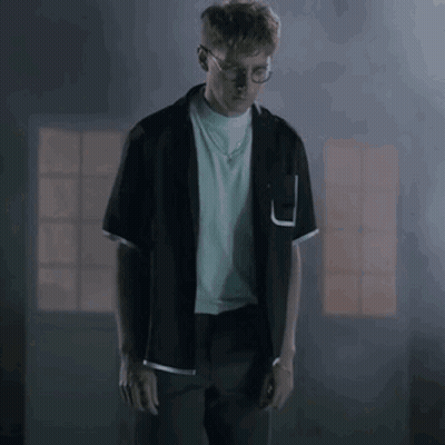 A Man In A Black Shirt And Glasses Is Standing In A Dark Room . GIF