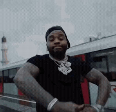 A Man In A Black Shirt And Hat Is Standing In Front Of A Bus . GIF