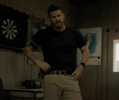 A Man In A Black Shirt And Khaki Pants Is Standing In Front Of A Dart Board In A Room . GIF