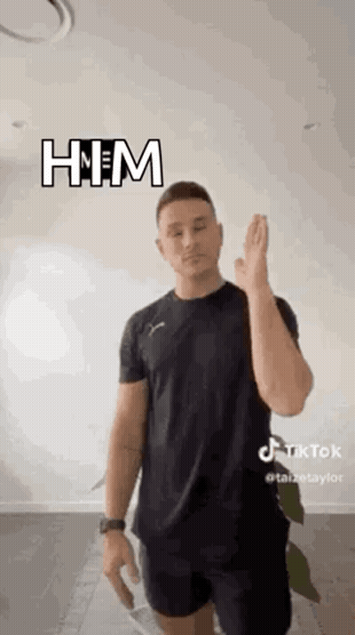 A Man In A Black Shirt And Shorts Is Standing In Front Of A White Wall And Giving A High Five . GIF