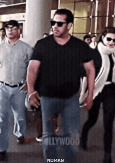 a man in a black shirt and sunglasses is walking with a woman in a white scarf behind him .