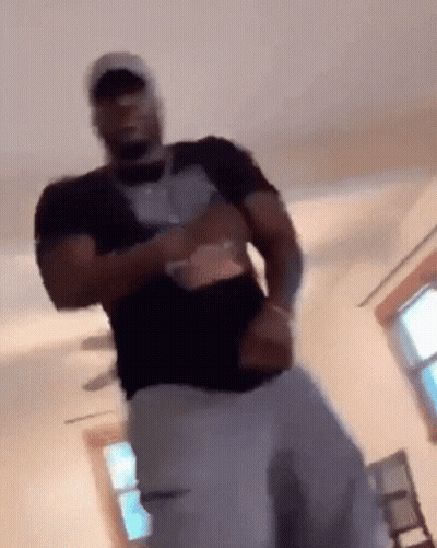 A Man In A Black Shirt And White Pants Is Standing In A Living Room . GIF