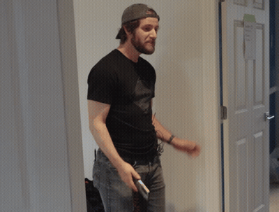 A Man In A Black Shirt Holds A Cell Phone In Front Of A Door GIF