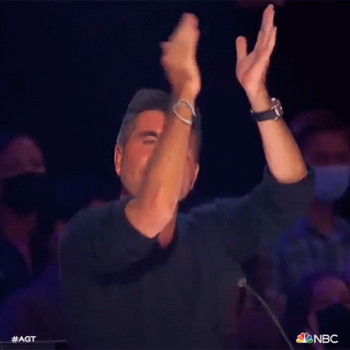 A Man In A Black Shirt Is Applauding While Sitting In Front Of A Crowd . GIF