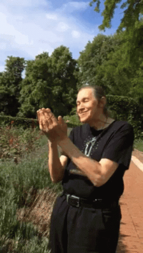 A Man In A Black Shirt Is Clapping His Hands In A Garden GIF