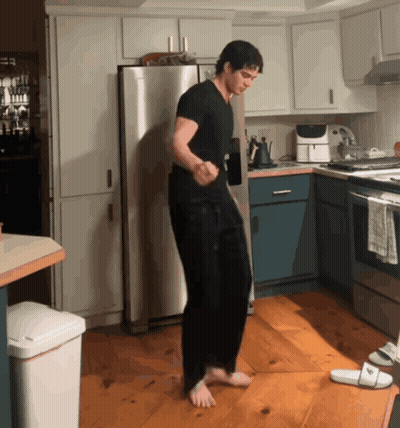 A Man In A Black Shirt Is Dancing In A Kitchen Next To A White Trash Can GIF