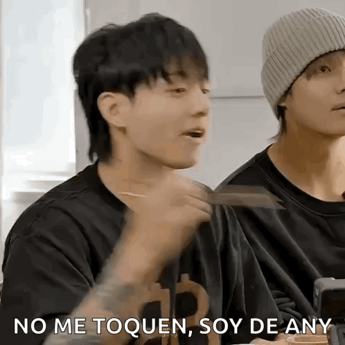 A Man In A Black Shirt Is Eating With Chopsticks While Another Man In A Beanie Looks On . GIF