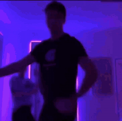 A Man In A Black Shirt Is Flexing His Muscles In A Room With Purple Lights . GIF